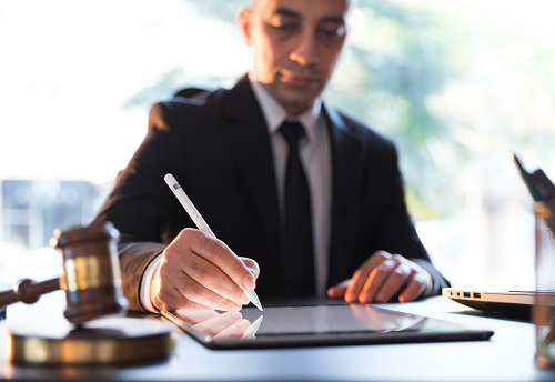 A lawyer at the courtroom is writing something on his tablet, wood hammer, glass of water and laptop can be seen.