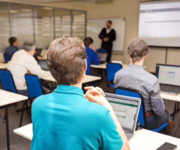 Rear view of trainees listening to their instructor and doing practical tasks on their laptops.